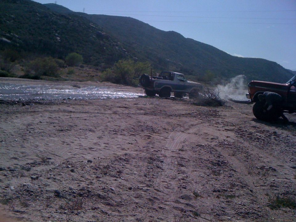 1978 Ford Bronco driving through shallow water, kicking up dust. Another truck is parked nearby