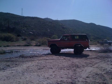 1978 Ford Bronco - A red 1978 Ford Bronco navigates a muddy, watery terrain with hills in the background.