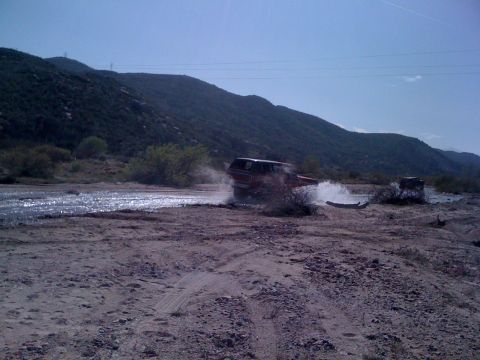1978 Ford Bronco - A red SUV kicks up water as it drives through a stream bed in a rocky, arid landscape.