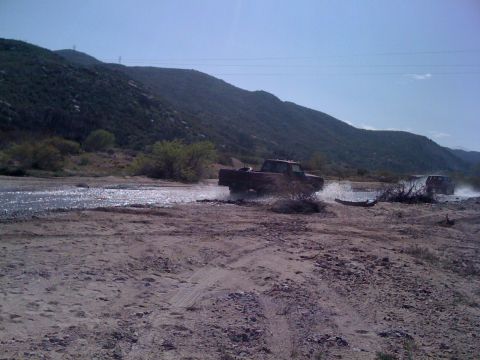 1978 Ford Bronco - A red pickup truck splashes through a shallow riverbed with a second vehicle visible in the distance.
