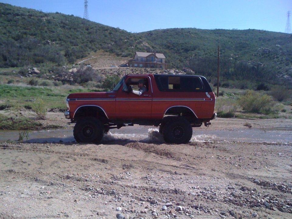 Red 1978 Ford Bronco driving through a shallow stream in a dry, hilly area