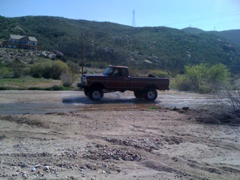 1978 Ford Bronco - A Ford F-Series pickup truck drives through a shallow stream in a dry, hilly landscape.