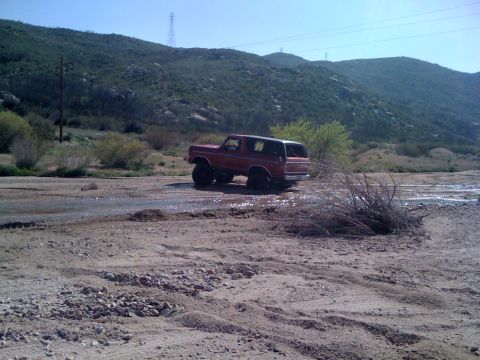 1978 Ford Bronco - Red 1978 Ford Bronco navigates a dry riverbed in a hilly, arid landscape.