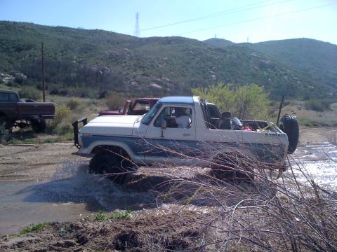 1978 Ford Bronco - A 1978 Ford Bronco navigates a muddy water crossing in a hilly, arid landscape.