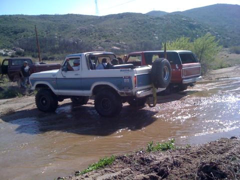 1978 Ford Bronco - Two off-road vehicles, including a 1978 Ford Bronco, navigate a muddy terrain under a clear sky.