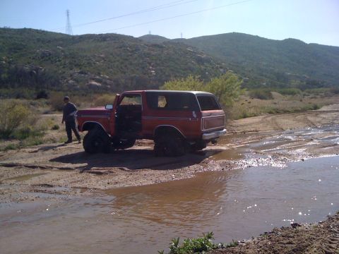 1978 Ford Bronco - A red 1978 Ford Bronco navigates a muddy water crossing with its driver's door ajar.