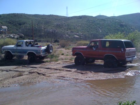 1978 Ford Bronco - Two 1978 Ford Broncos navigate a muddy off-road course under a clear blue sky.