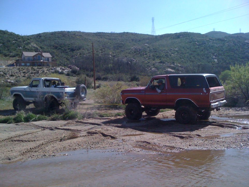 Two lifted Ford Broncos, one red and one blue/white, are off-roading in a muddy, hilly area. The red Bronco is being pulled by the blue/white one