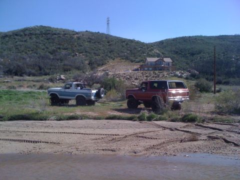 1978 Ford Bronco - Two classic Ford Broncos are parked in a rural landscape with a large house visible on the hillside.