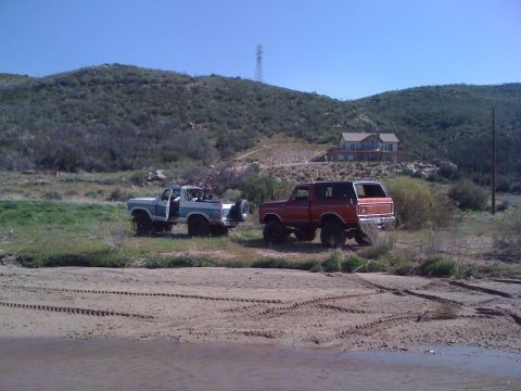 1978 Ford Bronco - Two 1978 Ford Broncos are parked in a rural landscape with a house and hills in the background.