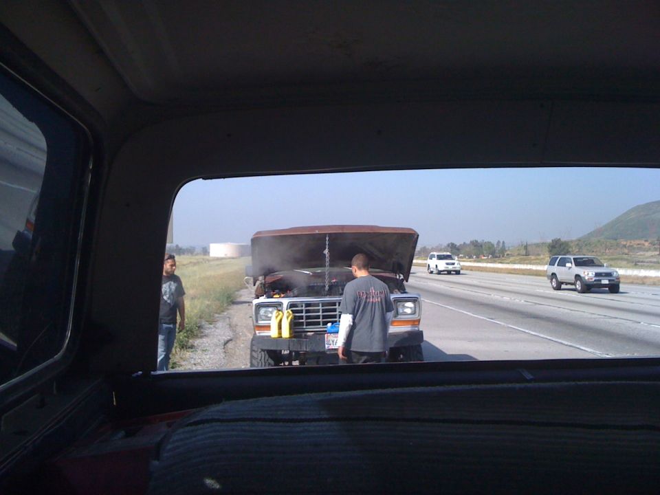 Truck with hood up on highway shoulder, steam coming out. Two people standing nearby. Other cars driving past