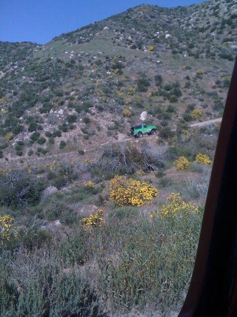1978 Ford Bronco - A green pickup truck navigates a dirt road through a dry, hilly terrain with scattered yellow wildflowers.