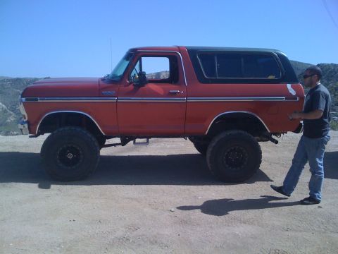 1978 Ford Bronco - A man stands next to a red, lifted 1978 Ford Bronco on a dirt road with hills in the background.