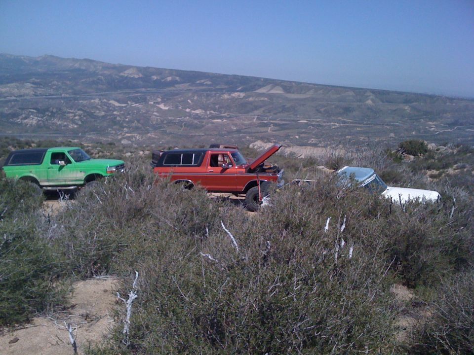 Orange 1978 Ford Bronco with hood open, parked next to a green Bronco and a white vehicle