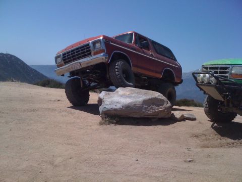 1978 Ford Bronco - Two off-road vehicles are seen on a dirt path with a large rock in the foreground.