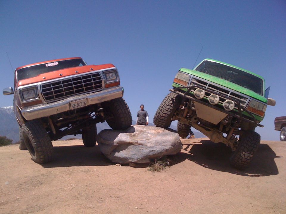 Orange and green lifted trucks are climbing over a large rock. One has a California license plate