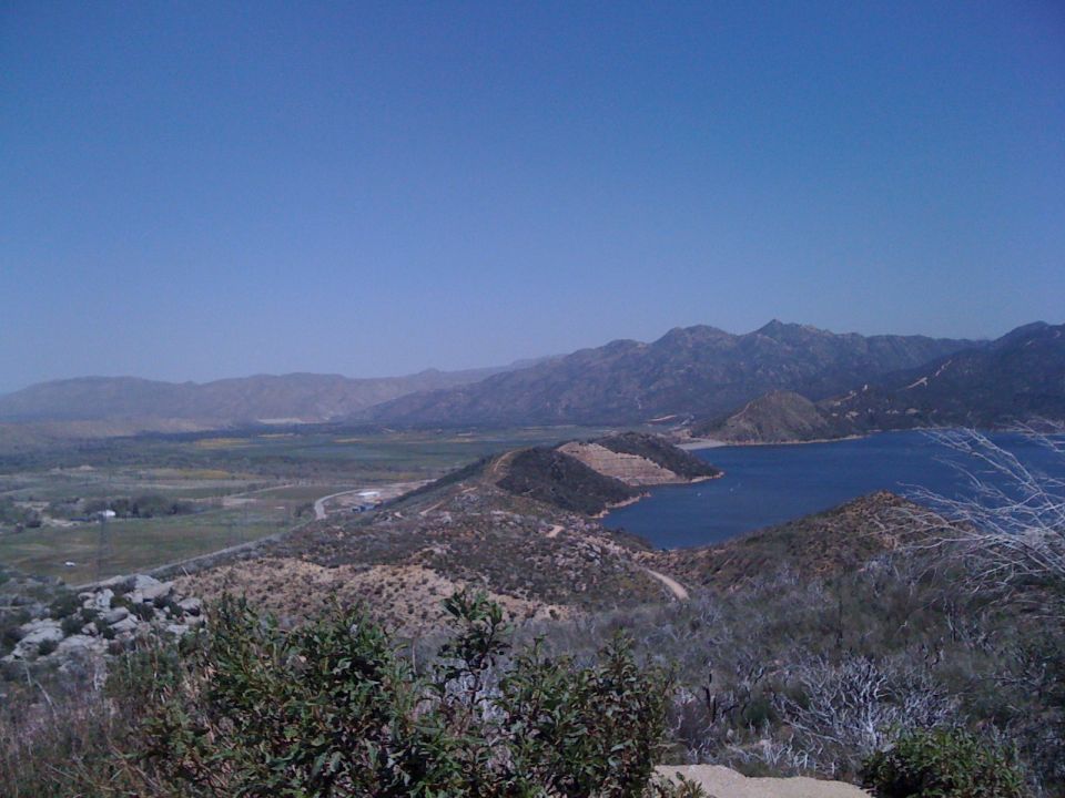 View of a blue lake surrounded by hills and mountains under a clear blue sky. Some vegetation is in the foreground