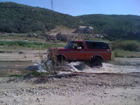 1978 Ford Bronco - A red 1978 Ford Bronco navigates a watery terrain, with a house visible on the distant hillside.