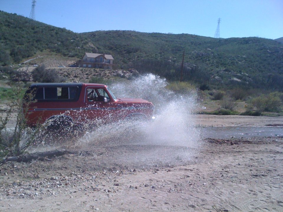 Red 1978 Ford Bronco driving through water, kicking up a large spray