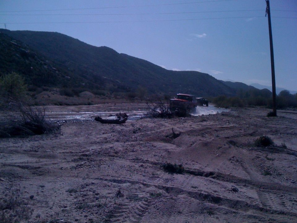 1978 Ford Bronco and another SUV driving through a dusty, dry riverbed with hills in the background