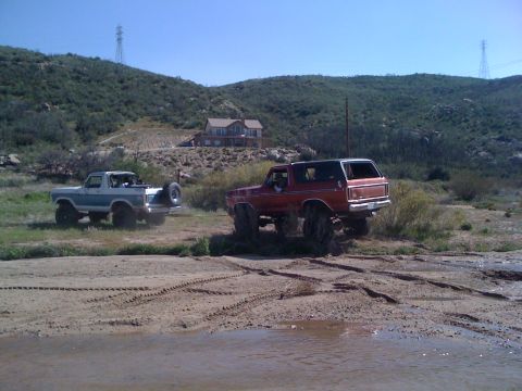 1978 Ford Bronco - Two Ford Broncos navigate a muddy off-road trail under a clear blue sky.