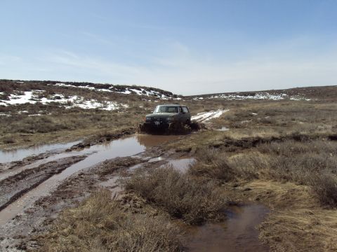 1989 Ford Bronco - A dark SUV navigates a muddy trail in a desolate, off-road environment under a clear blue sky.