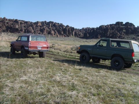 1989 Ford Bronco - Two SUVs, one green and one red, are parked on a grassy hill with rocky formations behind them.
