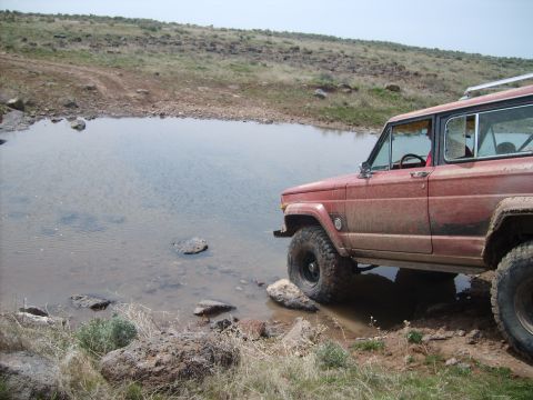1989 Ford Bronco - Jeep Cherokee XJ navigates off-road terrain near a water obstacle.