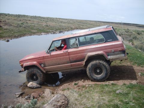 1989 Ford Bronco - A red Jeep Wagoneer navigates a rocky stream bed in a rural, grassy landscape.