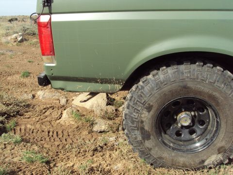 1989 Ford Bronco - The rear of a green SUV is shown on uneven, muddy terrain with rocks. A close-up of the off-road tire is visible.