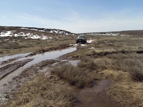 1989 Ford Bronco - 1989 Ford Bronco driving through a muddy, off-road trail with snow patches on the hills.