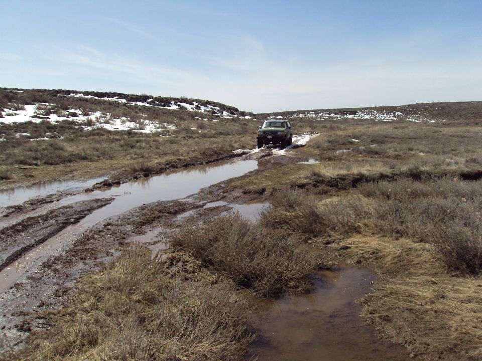 1989 Ford Bronco navigating a muddy track with puddles and sparse vegetation