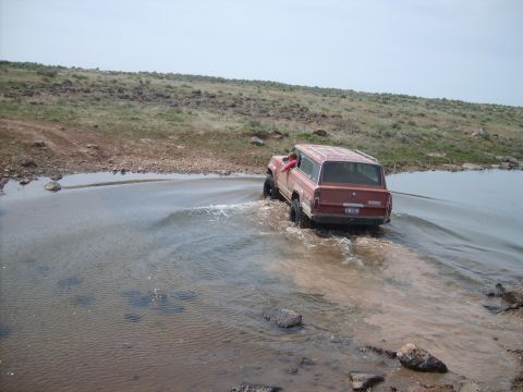 1989 Ford Bronco - A red Jeep Grand Wagoneer navigates through a shallow body of water on a dirt path.