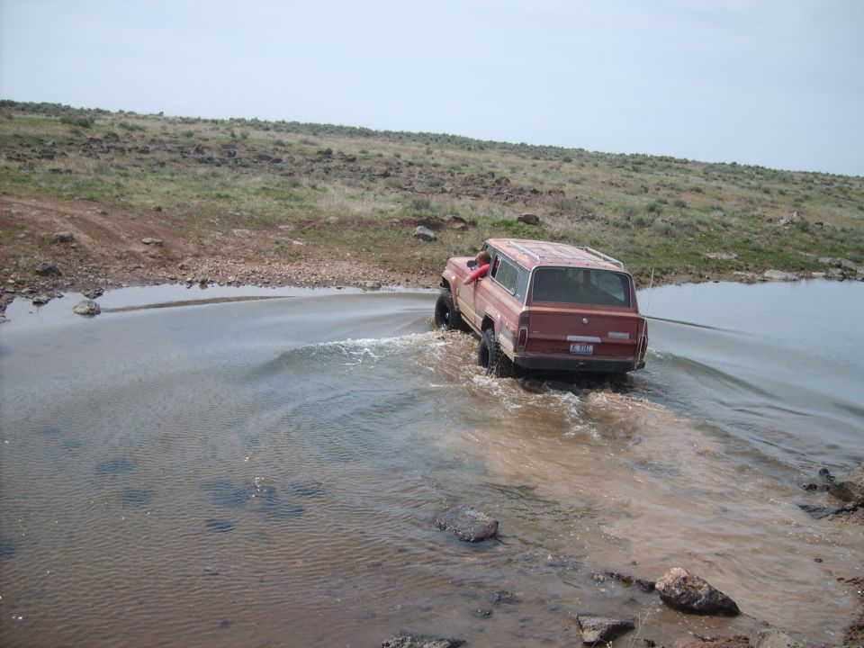 Red Jeep Grand Wagoneer driving through shallow water on a dirt road
