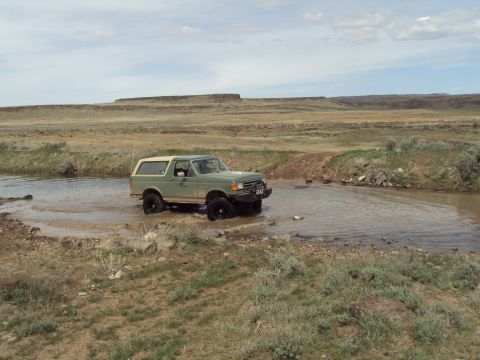 1989 Ford Bronco - A 1989 Ford Bronco navigates a shallow body of water in a dry, hilly landscape under a cloudy sky.