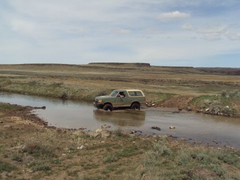 1989 Ford Bronco - A green 1989 Ford Bronco navigates a shallow water crossing in a vast, arid landscape under a cloudy sky.