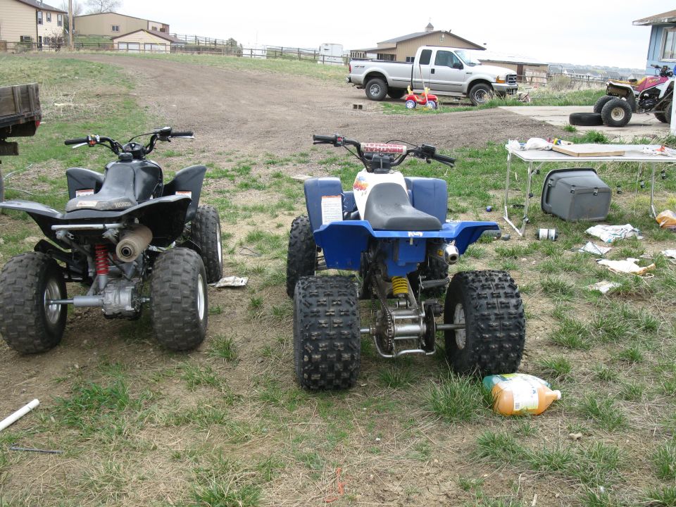 Two ATVs, one black and one blue, parked on a grassy area with a pickup truck and buildings in the background