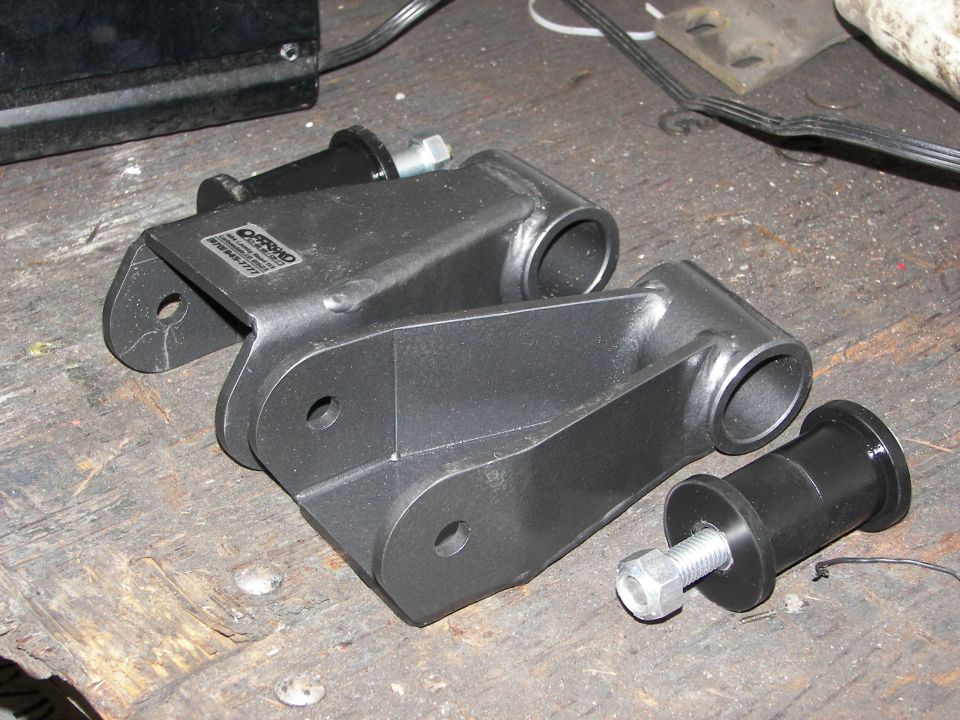 Pair of black metal suspension lift components with bushings and bolts on a wooden surface. One component has a label with text