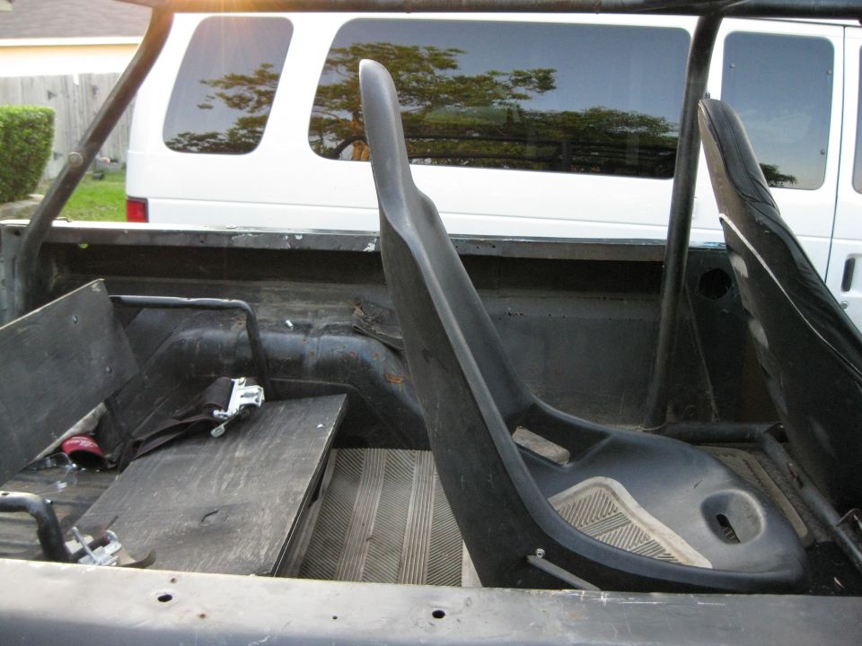 Interior view of a roll cage and racing seat, with a white van visible through the rear window