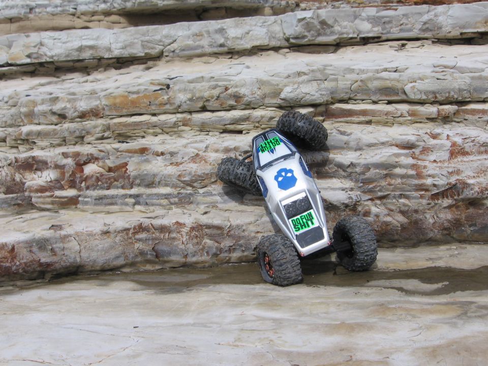 RC rock crawler on layered rocks. Text on body says 'DO FUN SHIT'
