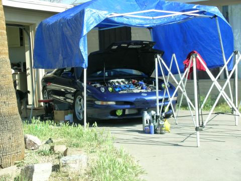 1995 Ford Probe - A blue coupe sits under a canopy with its hood open, revealing the engine bay and various bottles of fluids nearby.