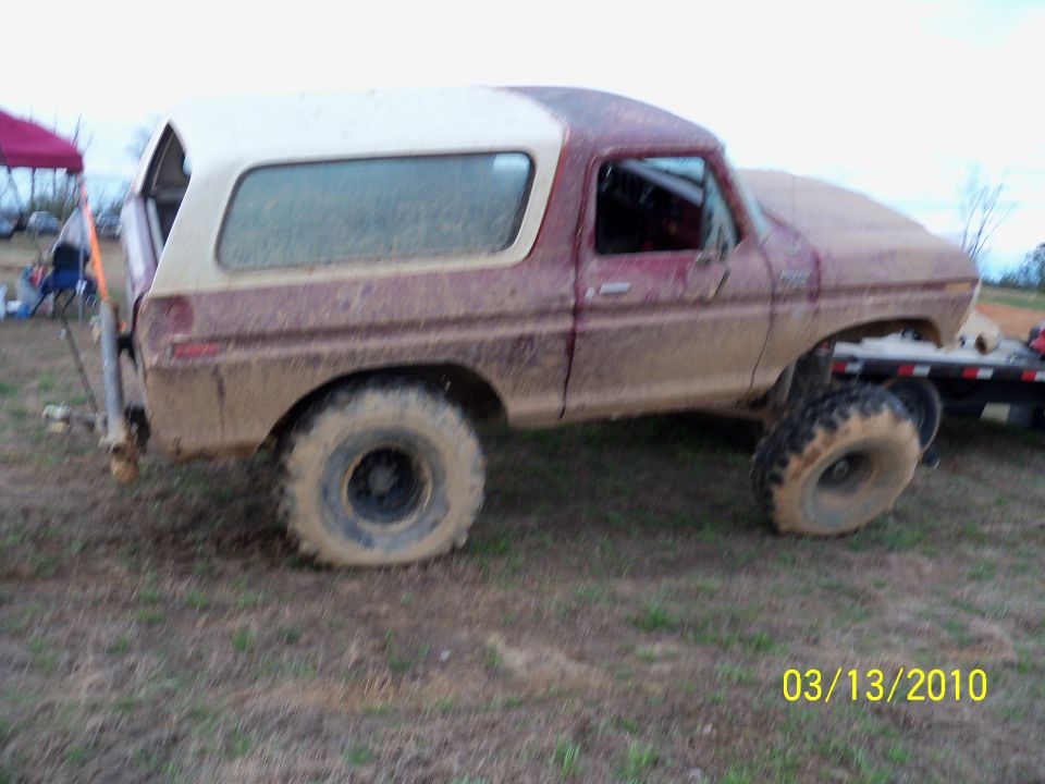 Mud-caked red Ford Bronco with white top, rear three-quarter view in a grassy field