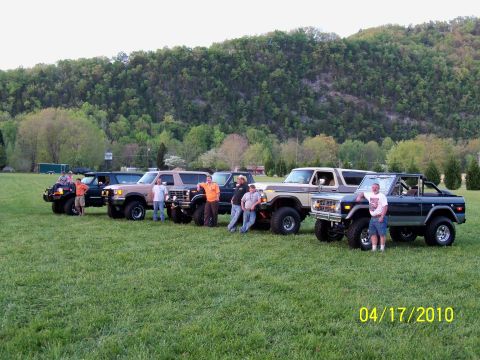 1979 Ford Bronco - A group of Ford Broncos are gathered on a grassy field with a wooded hill in the background.