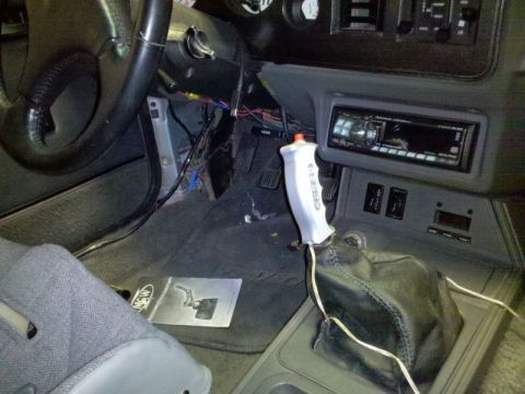 1986 Ford Mustang - Interior view of a car's center console and driver's footwell showing a white Hurst pistol-grip shifter handle mounted on a black leather s…