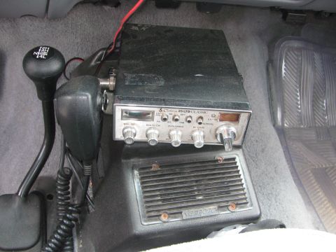 1996 Ford C-350 (Centurion Conversions) - Cobra 25 LTD Classic CB radio mounted on the transmission hump inside a truck cab, its handheld mic and coiled cord draped beside it and a…