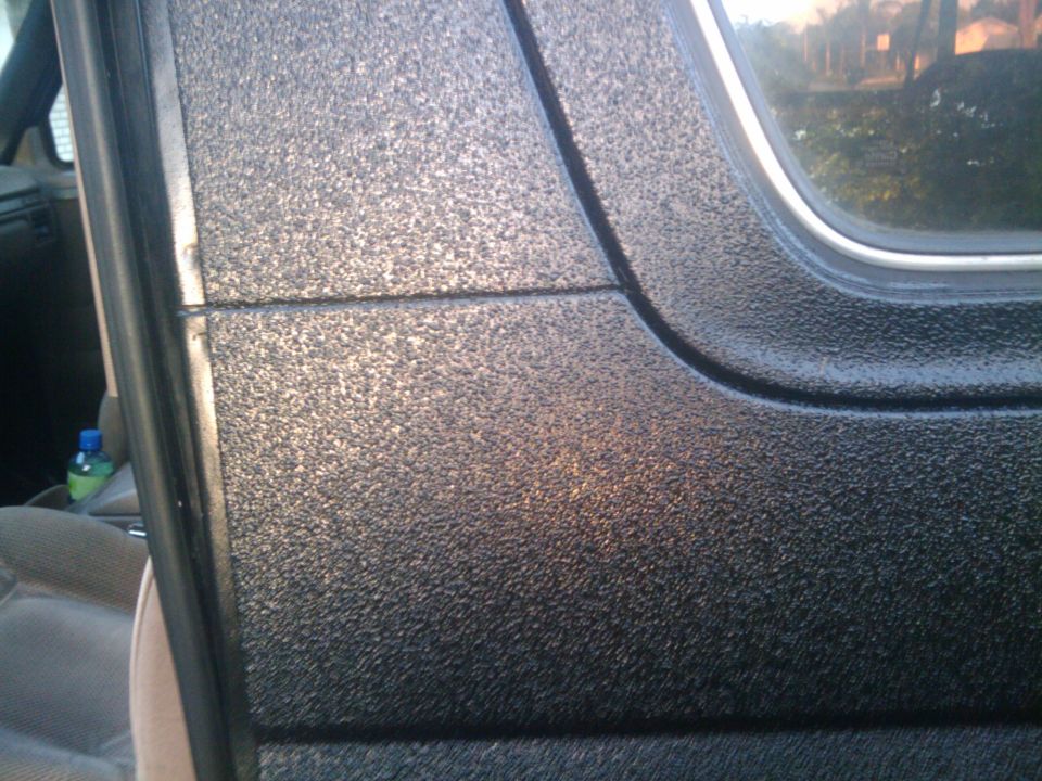 Close-up of textured black paint on a vehicle's door panel, showing interior seat and a bottle in the background