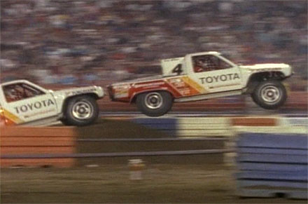 1986 Ford Bronco - Two Toyota trucks compete in a race, with one airborne over obstacles.