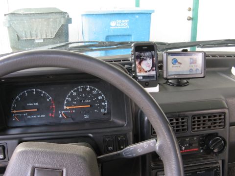 1991 Ford Festiva - The interior of a car shows a dashboard with a steering wheel, speedometer, and a mounted iPhone and Garmin GPS device.