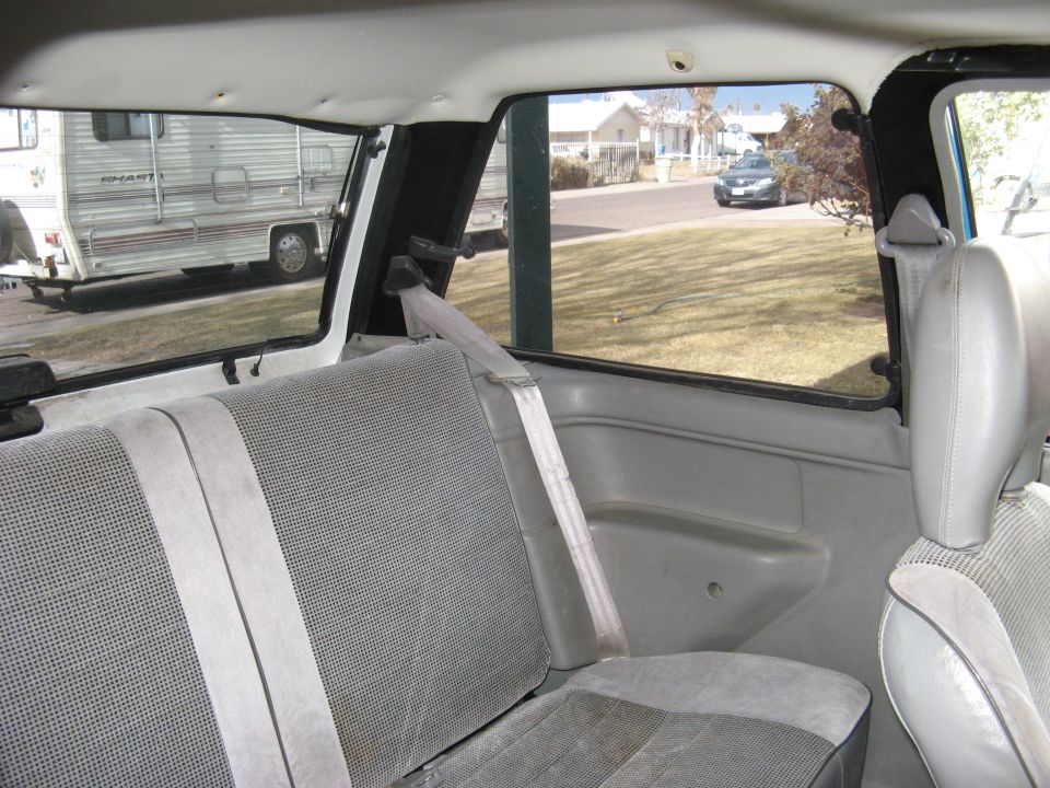 Interior view of a car showing rear seats, seatbelts, and a window looking out onto a street with houses and an RV parked nearby