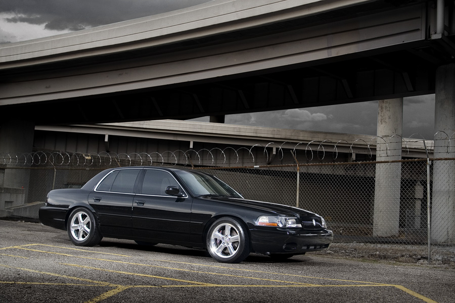 Black Mercury Marauder sedan front 3/4 under a concrete overpass with razor-wire fence